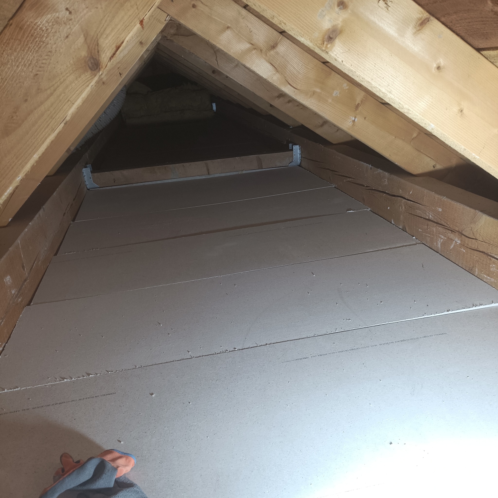 Wykończenie poddasza płytami gipsowymi, widoczne drewniane elementy konstrukcyjne dachu i fragment rękawicy roboczej w dolnym rogu kadru.