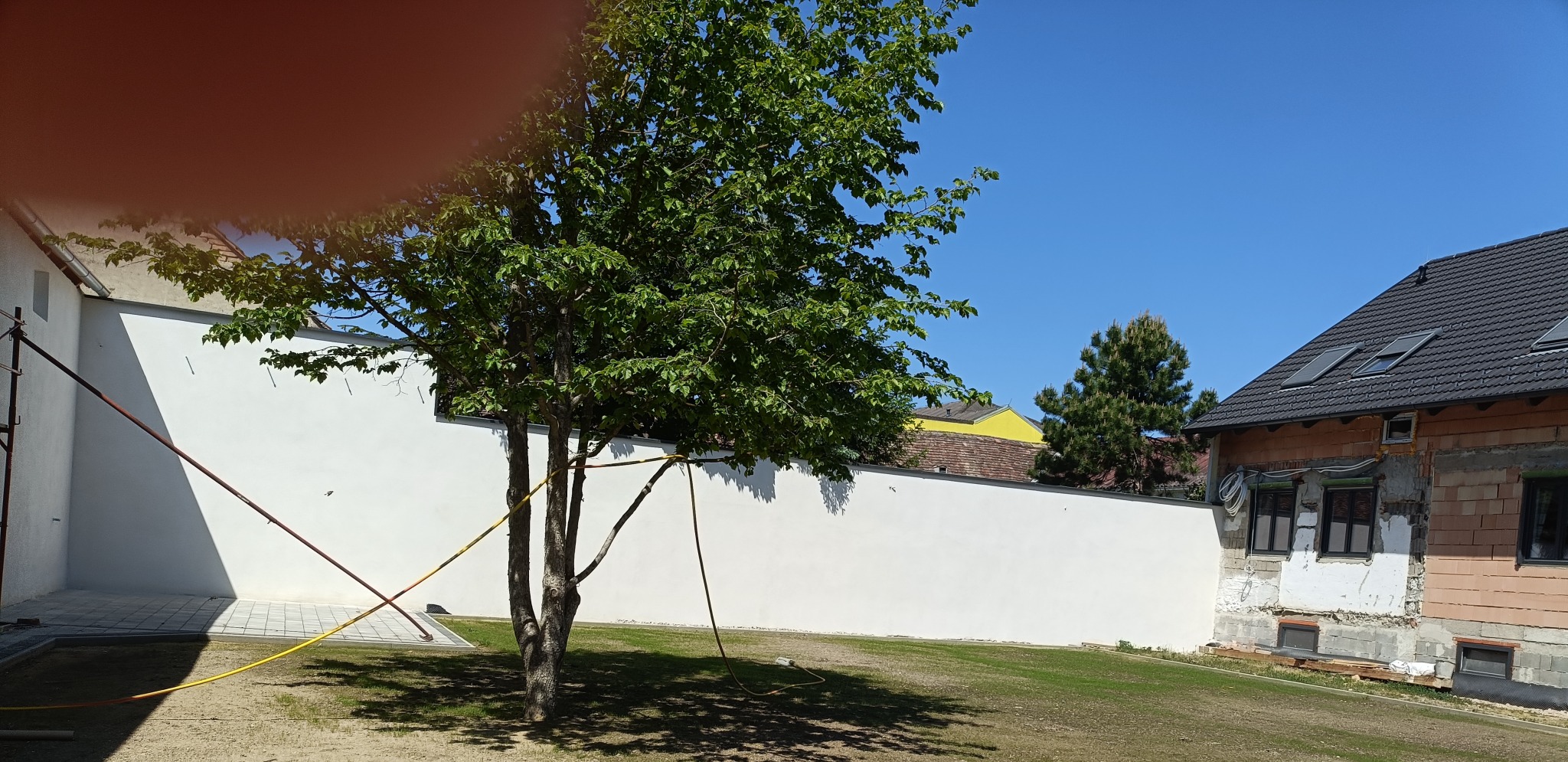 Biała, gładka ściana z rusztowaniem w tle. Fragment domu w trakcie budowy z oknami i dachem. Drzewo na pierwszym planie. Błękitne niebo.