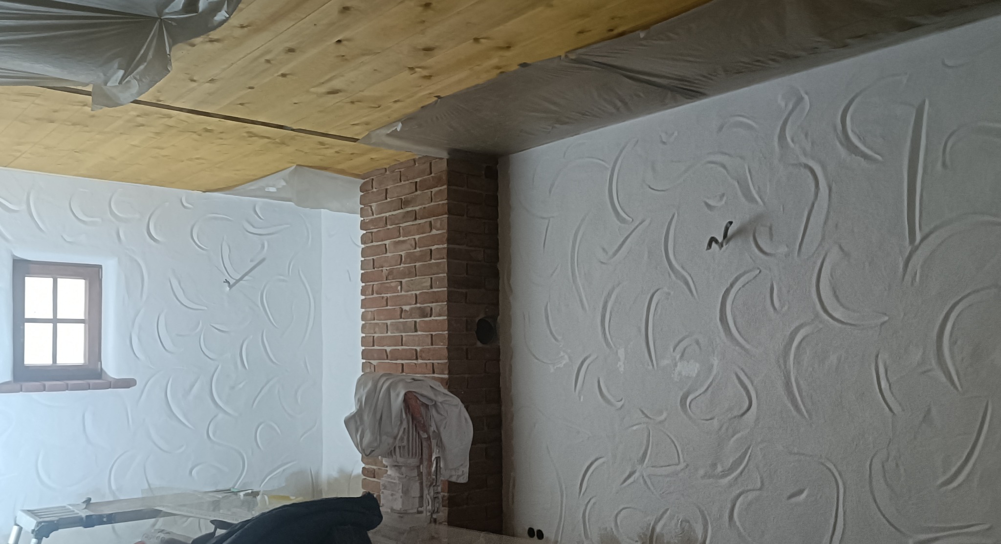 Wnętrze po tynkowaniu dekoracyjnym: białe ściany z subtelnym wzorem przypominającym fale, ceglany komin, drewniany strop i okno. Widoczne zabezpieczenia folią malarską.