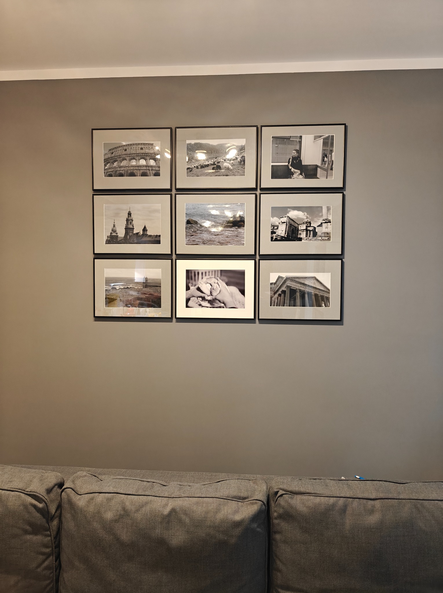 Ściana w szarym odcieniu z galerią dziewięciu czarno-białych, oprawionych zdjęć. Na dole widoczny fragment szarej sofy.