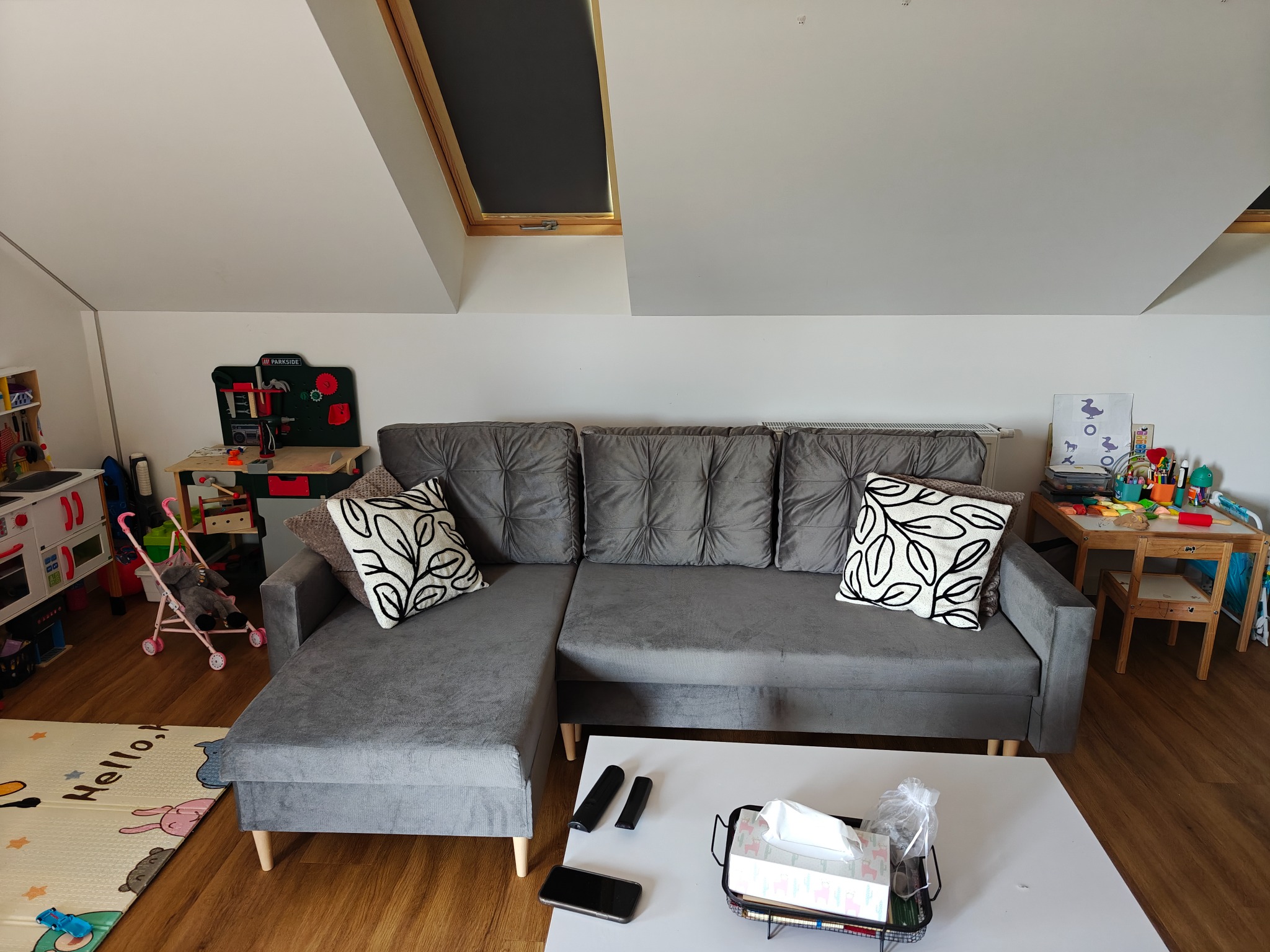 Szary narożnik z poduszkami w pokoju na poddaszu z oknem dachowym. W tle zabawki dziecięce i drewniana podłoga.