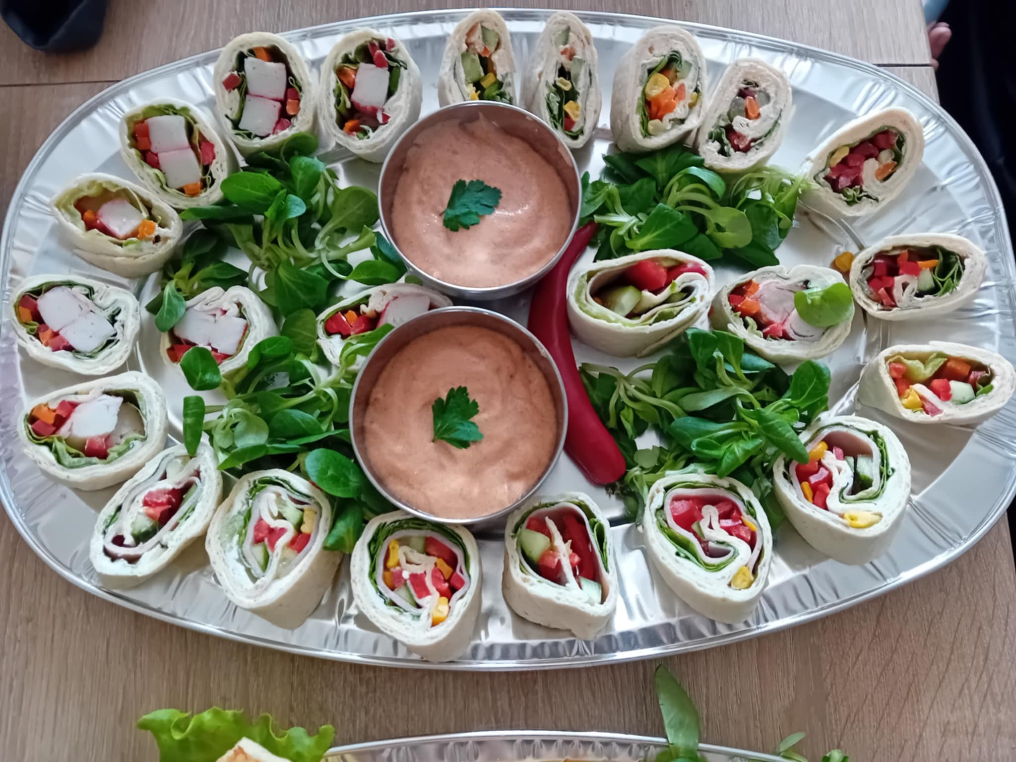 Srebrna taca z roladkami z tortilli z warzywami, sałatą i sosem, udekorowana świeżymi ziołami. Idealne jako przekąska na imprezę firmową.