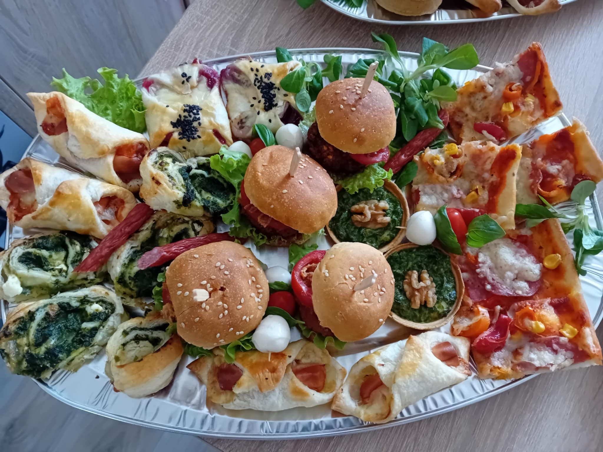 Srebrna patera z różnorodnymi przekąskami: mini burgery, pizza, roladki ze szpinakiem i ciastem francuskim, sałatki. Idealne na catering firmowy lub imprezę okolicznościową.