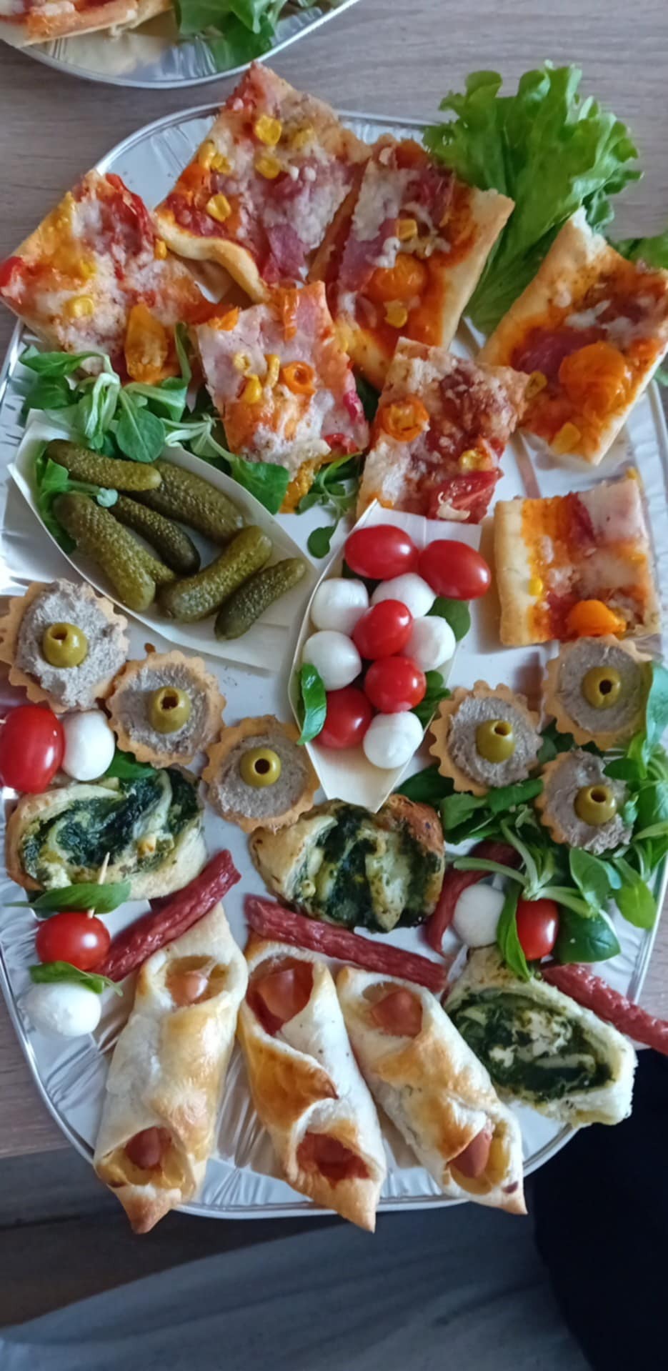 Elegancka taca z różnorodnymi przekąskami: pizza, tartaletki z pastą, koreczki caprese, roladki z ciasta francuskiego i parówki, ogórki konserwowe, tworząc apetyczny zestaw.