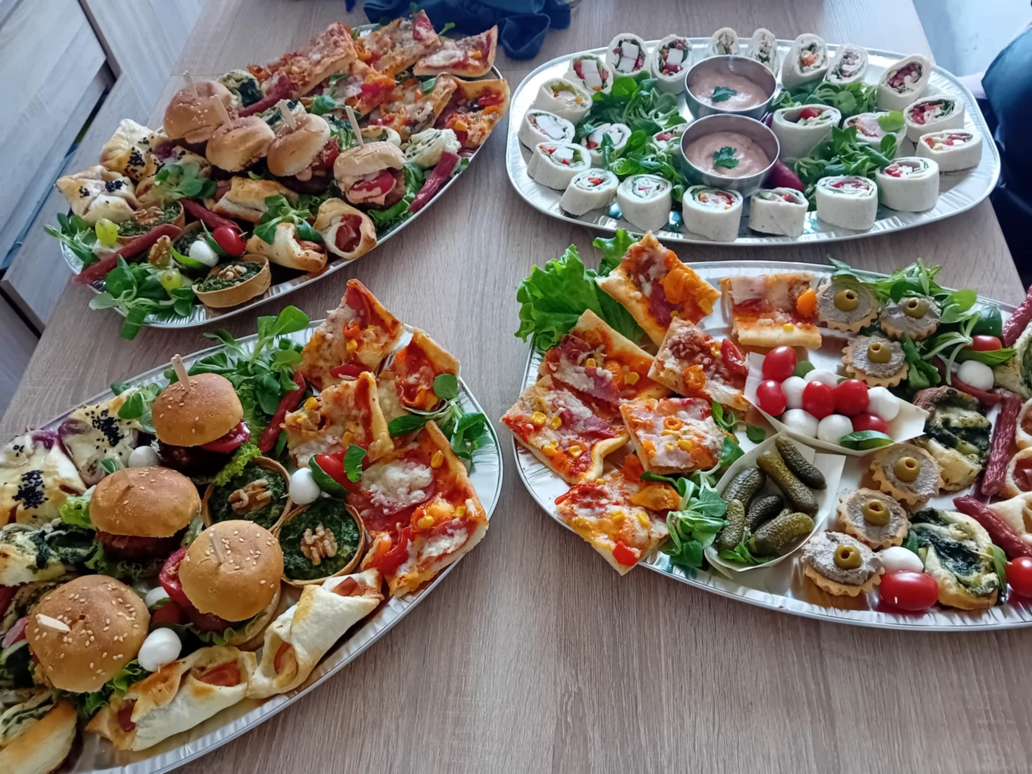 Bogato zastawiony stół z czterema paterami różnorodnych przekąsek: mini pizze, burgery, roladki, tartaletki i koreczki, udekorowane świeżymi warzywami. Idealne na przyjęcie.