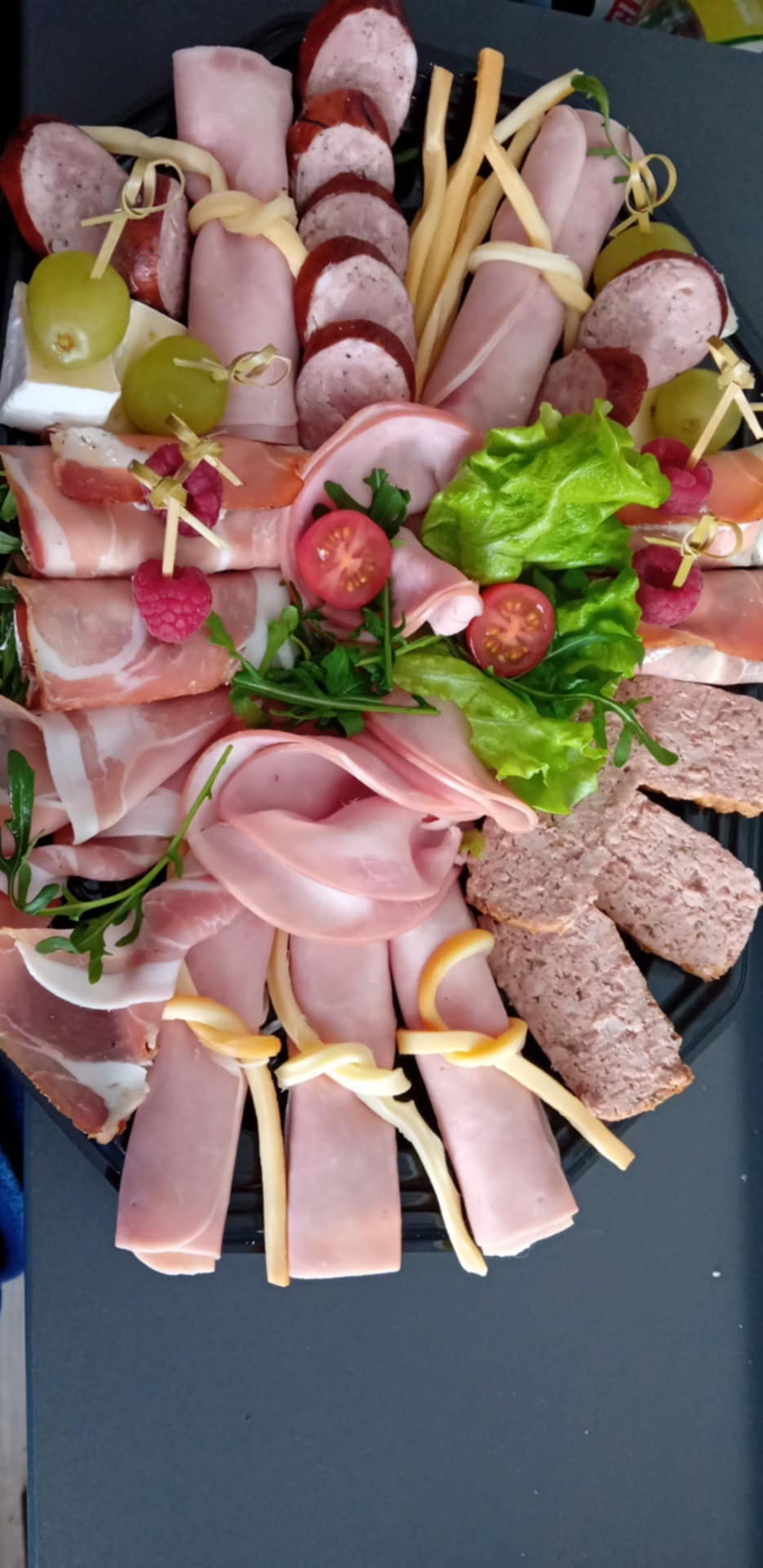 Dekoracyjna taca z wędlinami, serami i owocami, idealna na przyjęcie. Wędliny zwinięte w rulony, sery z winogronami, całość udekorowana zieleniną i pomidorkami.