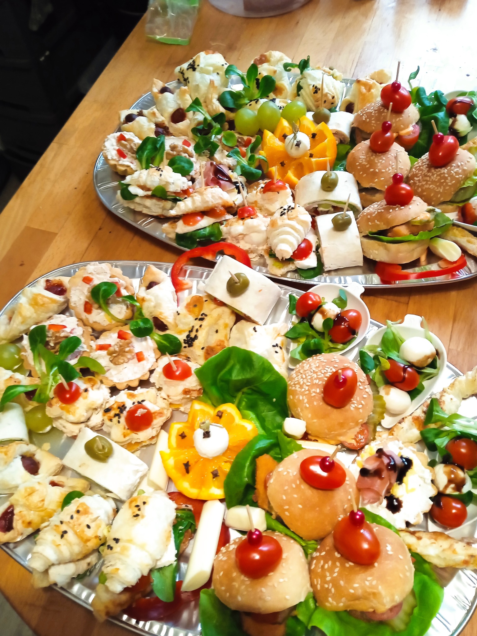 Dwie srebrne tace z różnorodnymi, apetycznymi przekąskami: mini burgery z sezamem, tartaletki, roladki z szynką, koreczki z pomidorkami i mozzarellą, dekoracje z pomarańczy.
