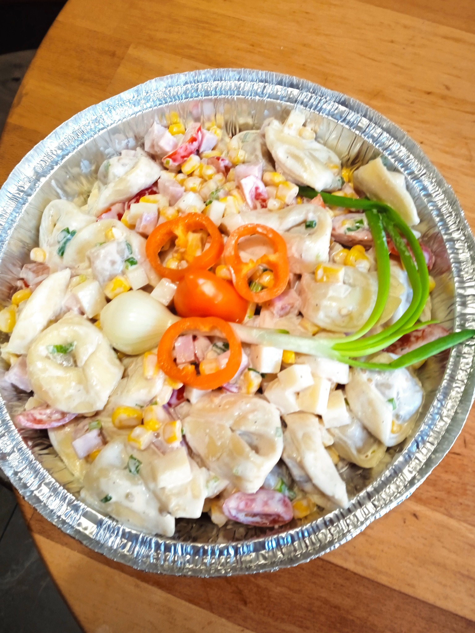 Sałatka z tortellini, kukurydzą, szynką, serem i warzywami w aluminiowej misce na drewnianym stole. Dekoracja z krążków papryki i szczypiorku.