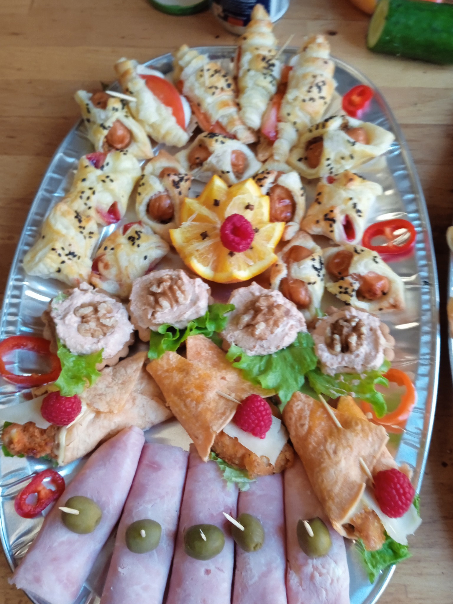 Srebrna patera z różnorodnymi przekąskami: roladki z szynki z oliwkami, tartaletki z pastą, mini tortille z malinami i ciastka francuskie z parówkami.