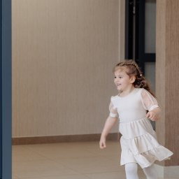 FotoDronik - Uśmiechnięta dziewczynka w jasnej sukience biegnie wzdłuż korytarza z beżowymi ścianami i podłogą z płytek. Widoczny fragment drzwi i futryny.