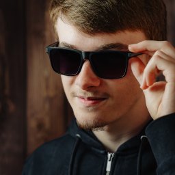 FotoDronik - Młody mężczyzna z rudymi włosami, w czarnej bluzie, przymierza czarne okulary na tle drewnianej ściany. Portret w zbliżeniu, oświetlenie podkreśla detale twarzy.