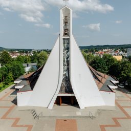 FotoDronik - Nowoczesny kościół z wieżą i dzwonami, widok z góry. Na pierwszym planie wzorzysty chodnik z kostki i zabytkowy samochód ślubny. W tle zabudowa miejska i błękitne niebo.