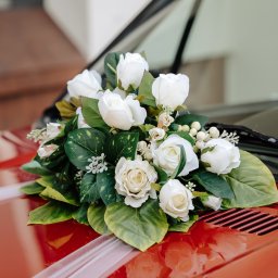 FotoDronik - Bukiet białych róż z zielonymi liśćmi leży na czerwonej masce samochodu z białymi wstążkami. Widoczne wycieraczki i odbicie w lakierze.