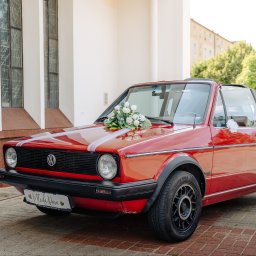 FotoDronik - Czerwony kabriolet VW Golf udekorowany kwiatami i wstążkami, tablica rejestracyjna z napisem 'Młoda Para', tło to budynek z oknami i drzewa.