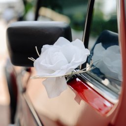 FotoDronik - Dekoracja z białego kwiatu na lusterku czerwonego samochodu. Detal z uroczystości, widoczne odbicie w szybie. Ujęcie z bliska.