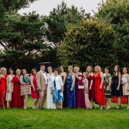 FotoDronik - Grupowe, plenerowe zdjęcie gości weselnych w różnorodnych, eleganckich strojach, z parą młodą w centrum, na tle zieleni drzew i trawnika.