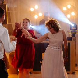 FotoDronik - Rozświetlona sala balowa, goście w ruchu podczas tańca, kobieta w sukni wieczorowej wskazuje kierunek, DJ w tle, światła punktowe tworzą klimat imprezy.