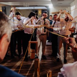 FotoDronik - Grupa mężczyzn w eleganckich strojach uczestniczy w grze z linami podczas uroczystości. W tle widoczne elementy dekoracyjne sali.