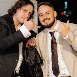 FotoDronik - Dwóch uśmiechniętych mężczyzn w garniturach pozuje, pokazując gest 'shaka'. Jeden w beżowym garniturze z brązowym krawatem, drugi w czarnym. Nocne oświetlenie w tle.