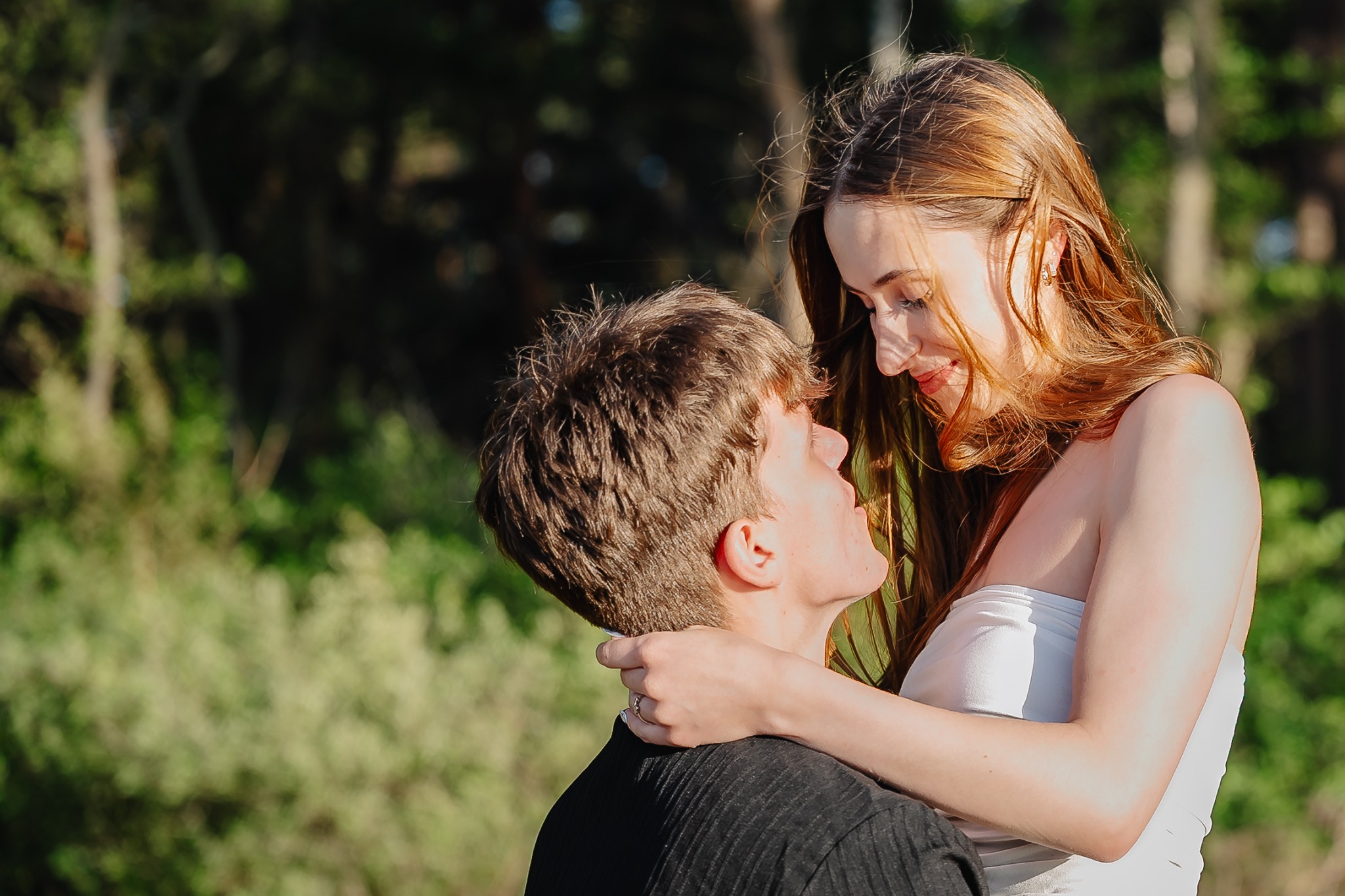 Zakochana para w plenerze: on w czarnej koszuli, ona w białej sukience, obejmują się, patrząc sobie w oczy na tle zieleni i drzew.