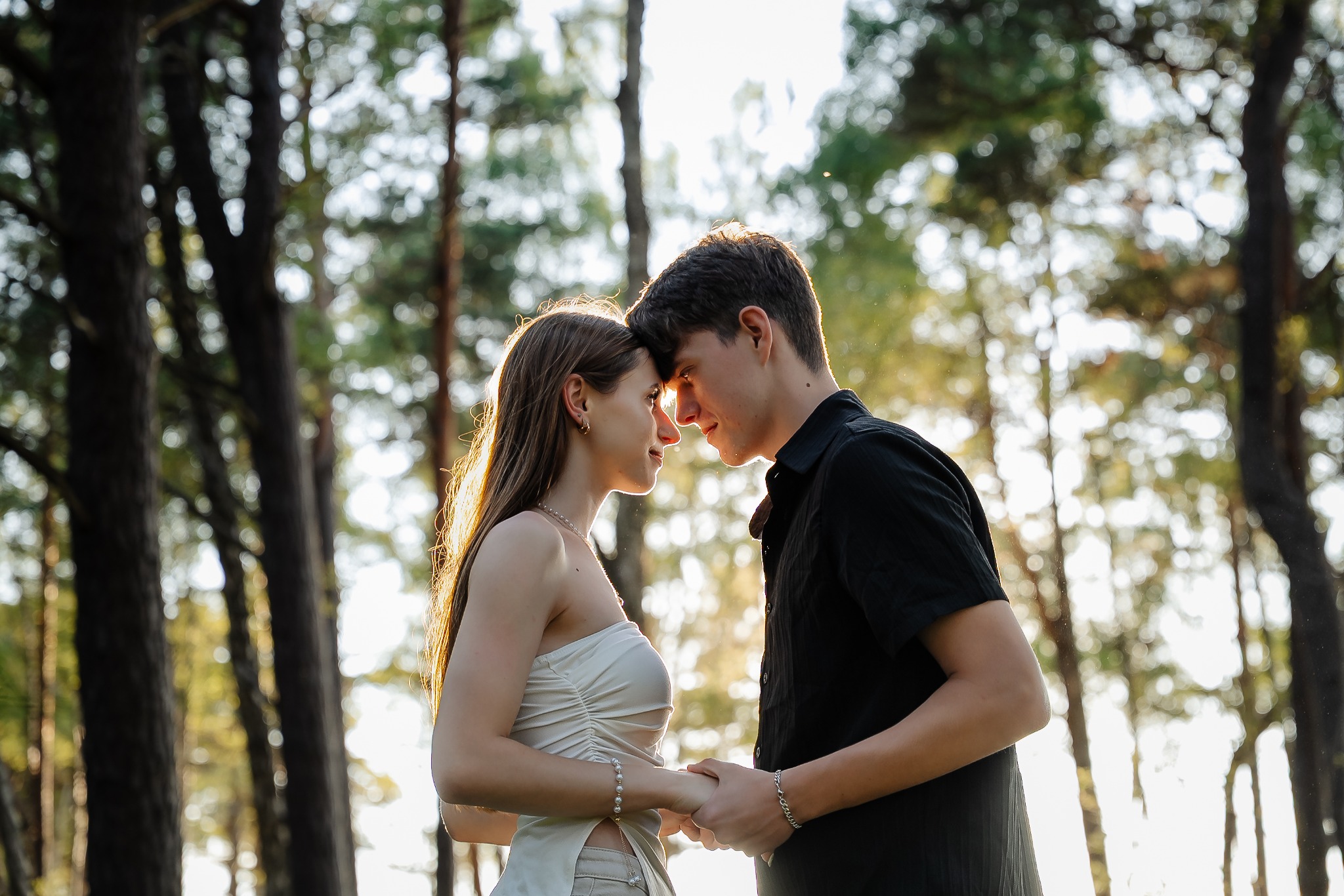 Zakochana para trzyma się za ręce, stykając się czołami na tle leśnych drzew w ciepłym świetle. Romantyczna sesja plenerowa.