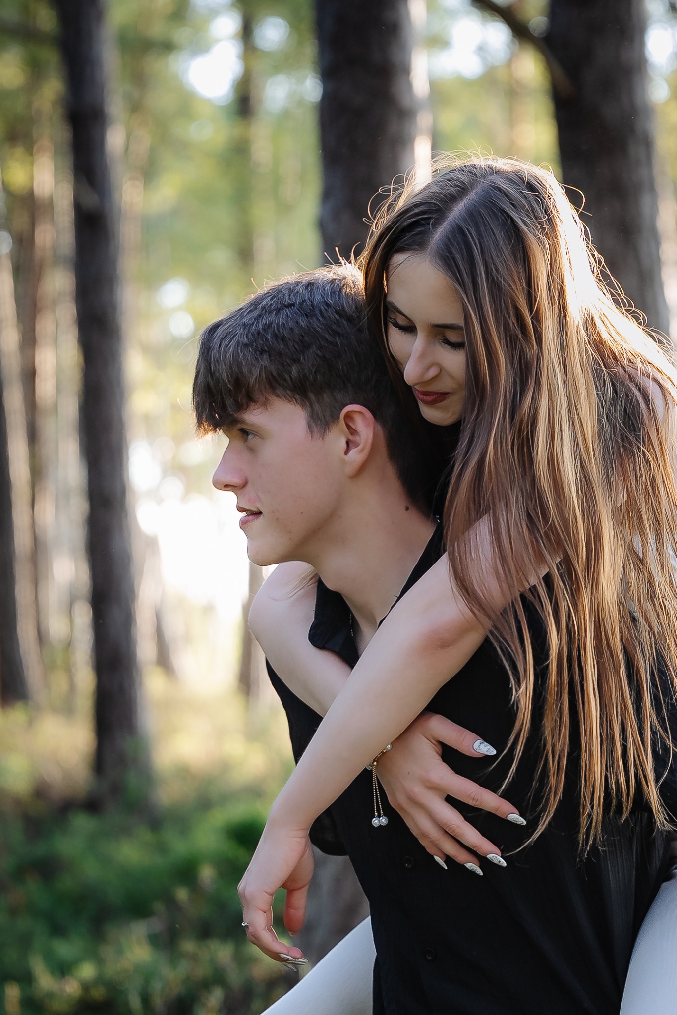 Młoda para w lesie: chłopak niesie dziewczynę na barana, ona obejmuje go, promienie słońca przebijają przez drzewa, tworząc ciepłą atmosferę. Uśmiechnięta dziewczyna z długimi włosami.