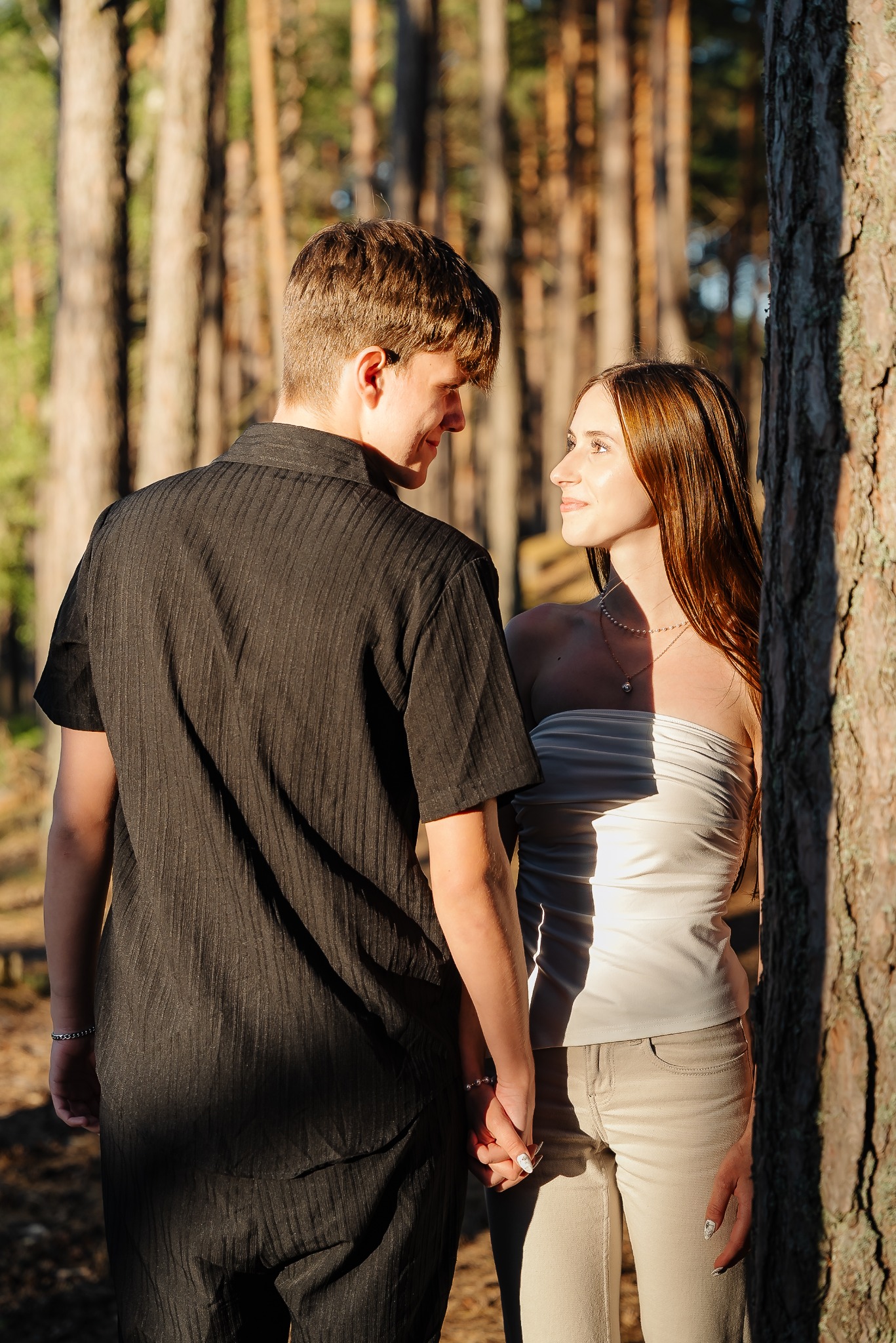 Para trzymająca się za ręce w lesie, ona w jasnej bluzce i spodniach, on w czarnej koszuli, oboje patrzą na siebie, ciepłe światło słoneczne.