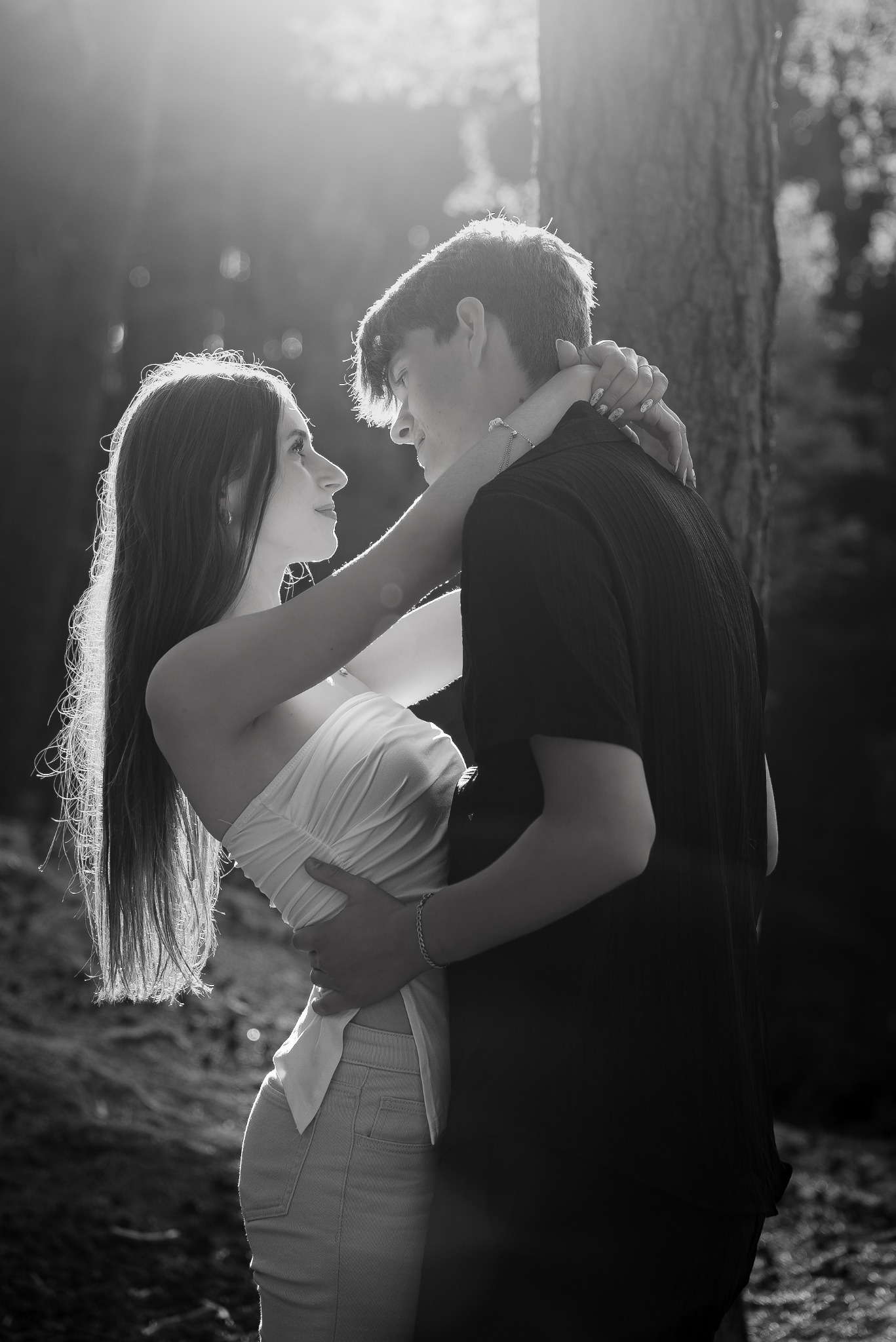 Czarno-białe, pełne czułości ujęcie obejmującej się pary w leśnej scenerii, z silnym światłem padającym zza pleców kobiety, podkreślającym jej sylwetkę.