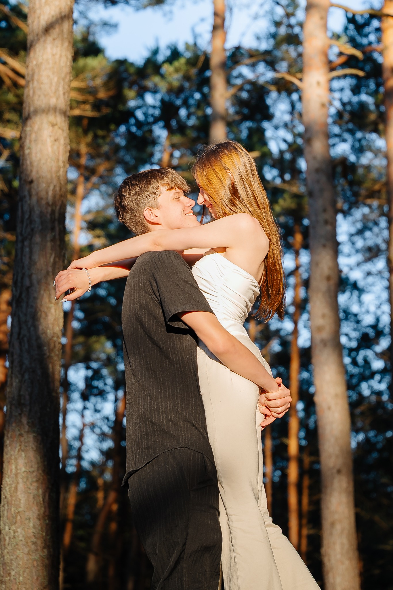Zakochana para w lesie: mężczyzna w czarnej koszuli podnosi kobietę w białym kombinezonie, patrząc sobie w oczy na tle drzew.