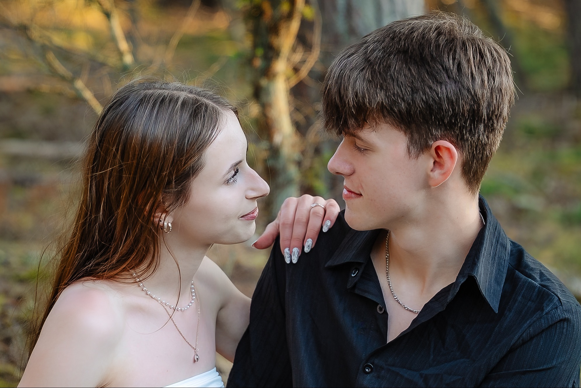 Romantyczna sesja narzeczeńska w plenerze: młoda para wpatruje się w siebie, on w czarnej koszuli, ona w białej sukience, delikatna biżuteria, naturalne światło.