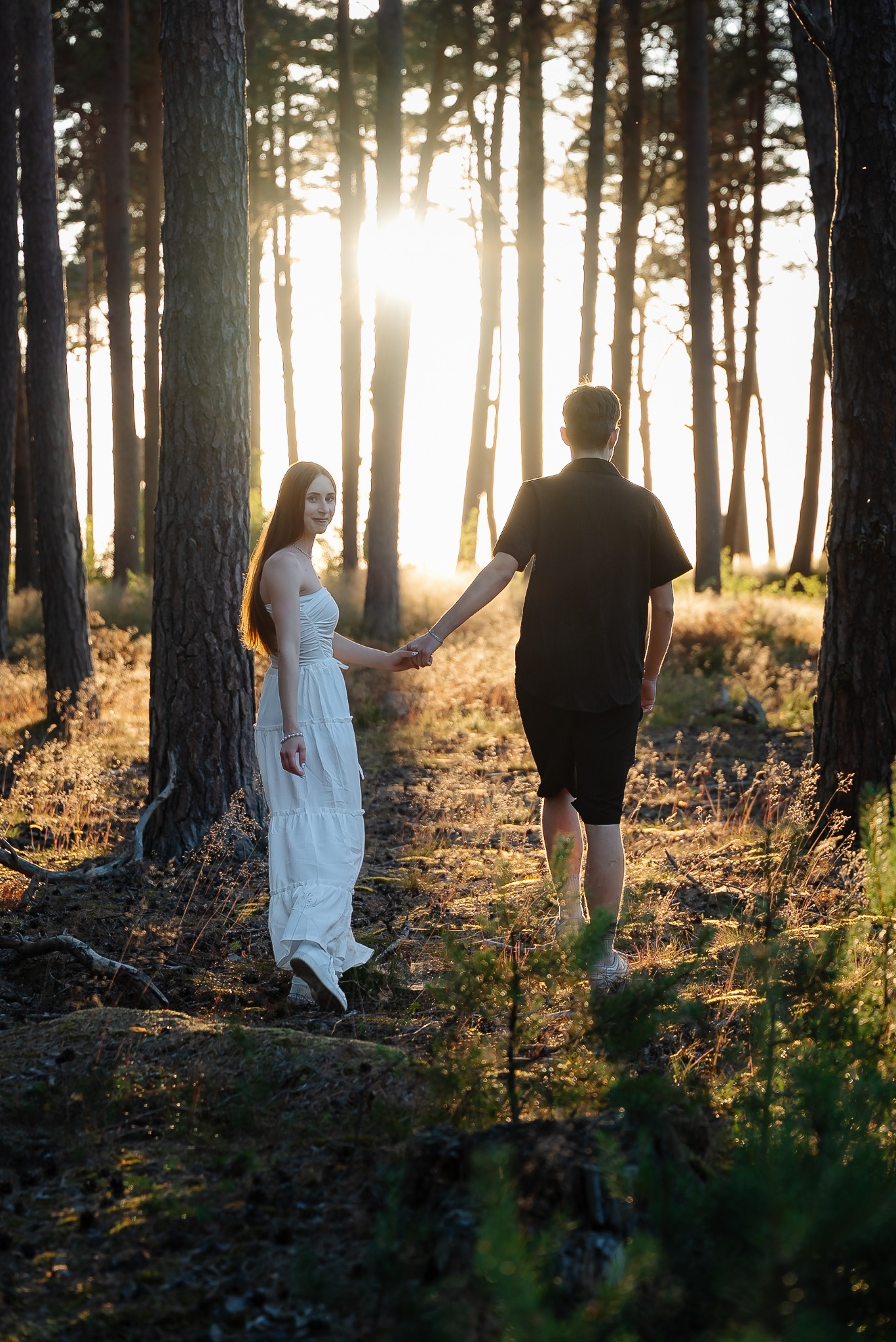 Para trzymająca się za ręce spaceruje po lesie w promieniach zachodzącego słońca. Kobieta w białej sukience spogląda w stronę kamery.