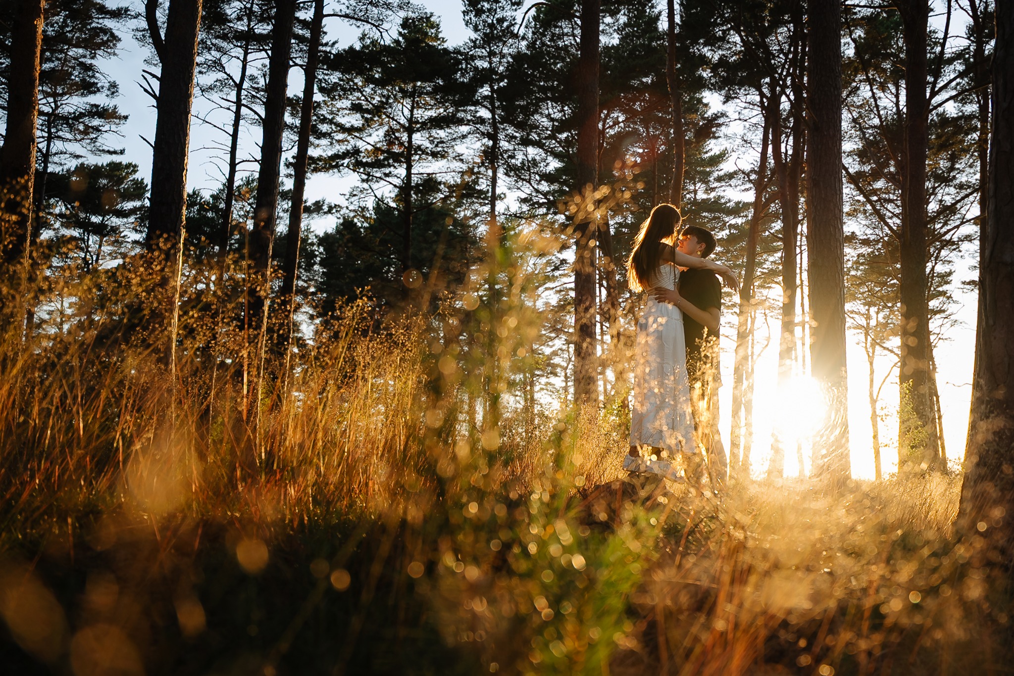 Zakochana para w objęciach w lesie o zachodzie słońca. Dziewczyna w białej sukience, chłopak w czarnej koszulce. Ciepłe światło przebija przez drzewa i trawy.