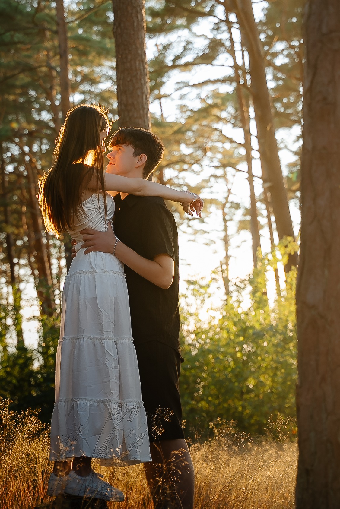 Zakochana para w leśnej scenerii, on obejmuje ją, promienie słońca przebijają przez drzewa, tworząc romantyczny nastrój. Sesja narzeczeńska w naturalnym otoczeniu.