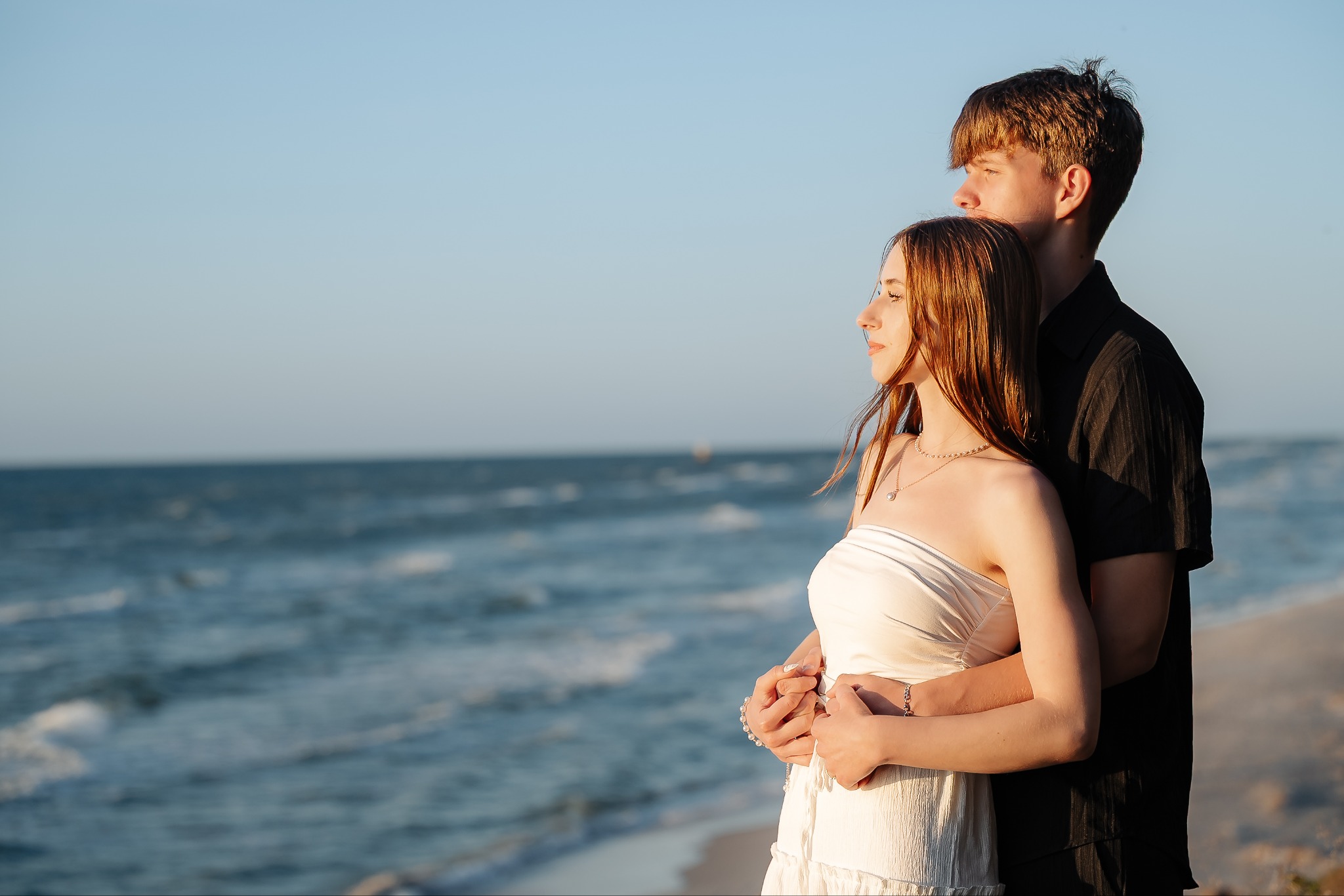 Zakochana para na plaży, obejmująca się, patrzy w dal na morze. Romantyczna sesja zdjęciowa w ciepłym, naturalnym świetle.