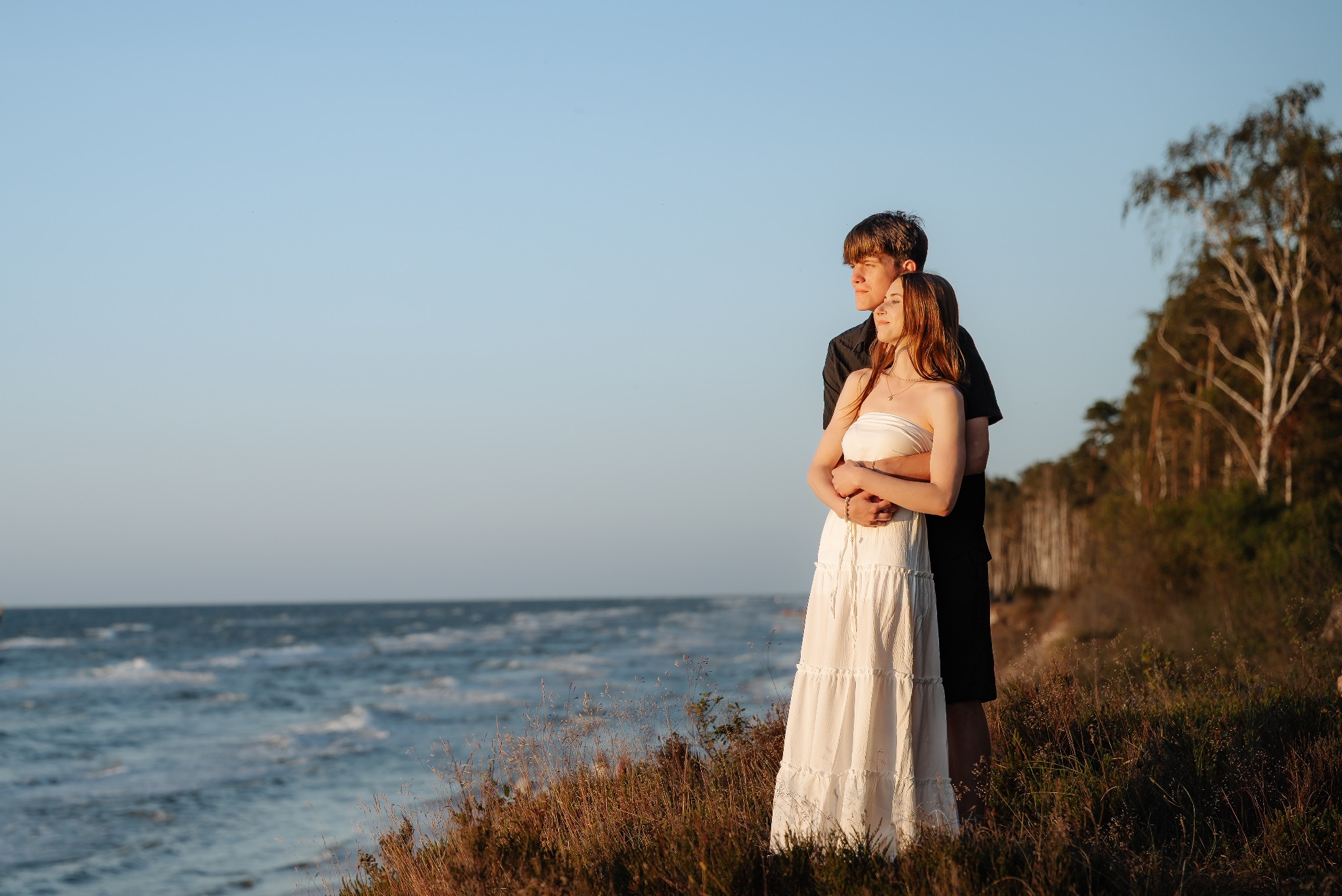 Zakochana para na tle morza w promieniach zachodzącego słońca. Kobieta w białej sukience, mężczyzna obejmuje ją od tyłu. Romantyczna sesja na klifie, naturalne światło.