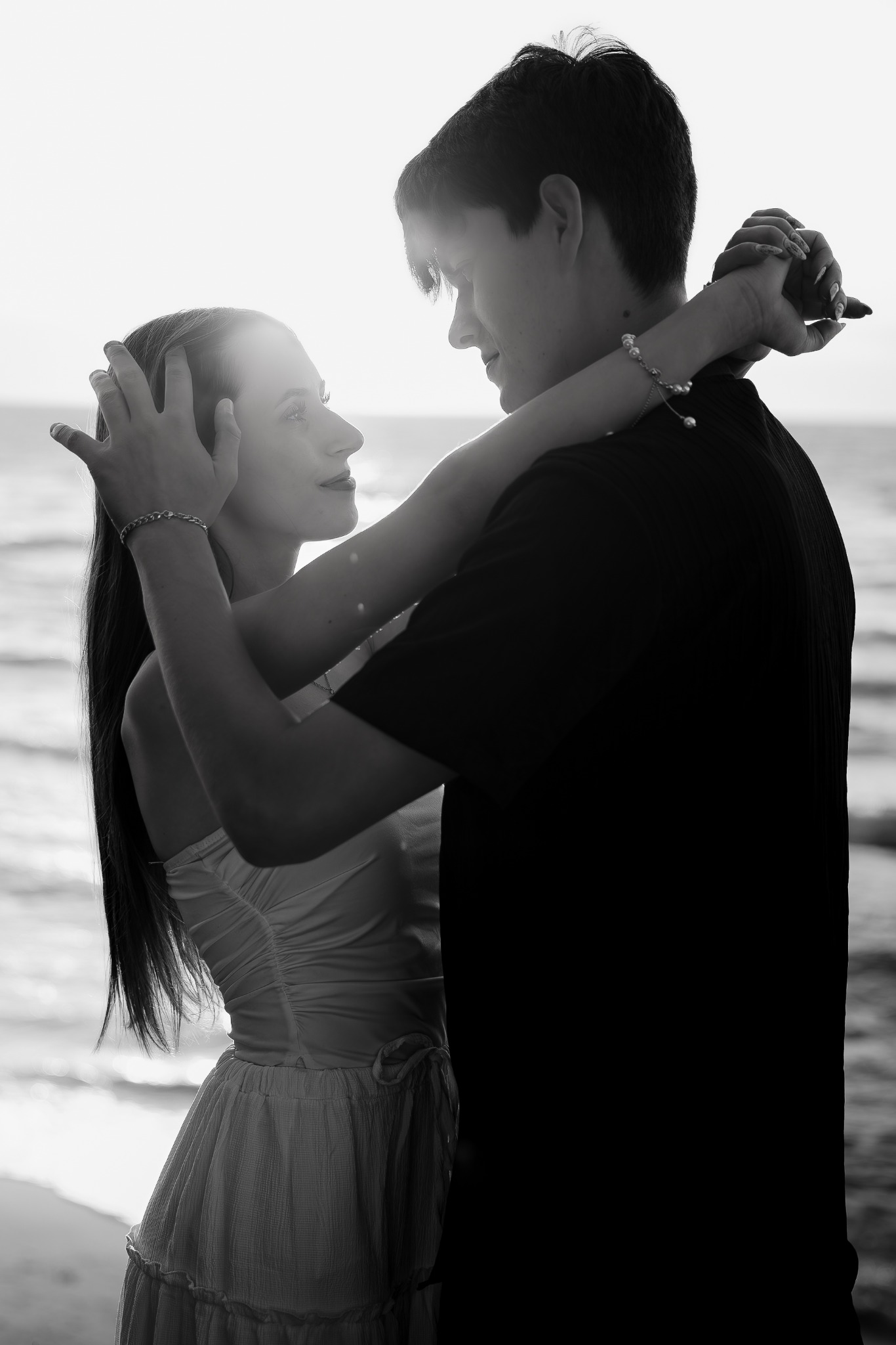 Czarno-białe zdjęcie zakochanej pary na plaży, kobieta obejmuje mężczyznę, patrząc mu w oczy. Delikatne światło podkreśla bliskość i intymność momentu.