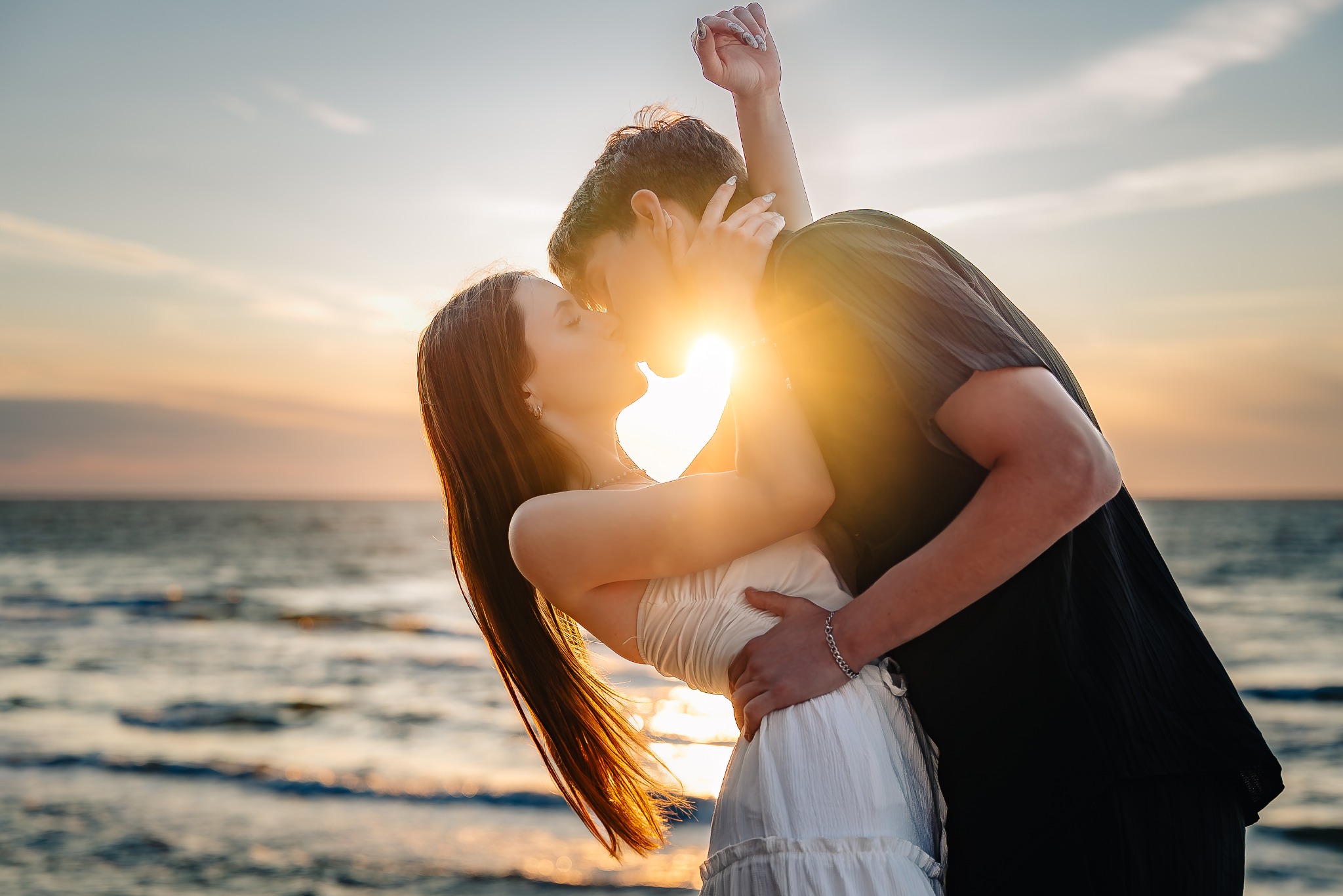 Zakochana para na plaży podczas zachodu słońca. Kobieta w białej sukience, mężczyzna w czarnej koszuli. Silne światło słoneczne tworzy romantyczną atmosferę, podkreślając bliskość.