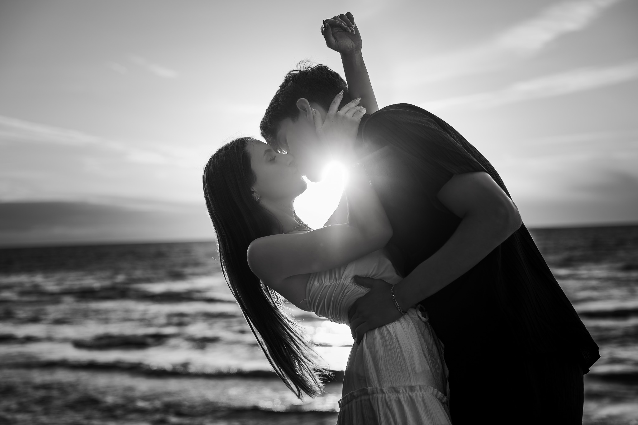 Czarno-białe zdjęcie zakochanej pary na tle morza i słońca. Kobieta w jasnej sukience, mężczyzna w ciemnej koszuli, oboje się całują, a mężczyzna trzyma rękę uniesioną w górę.