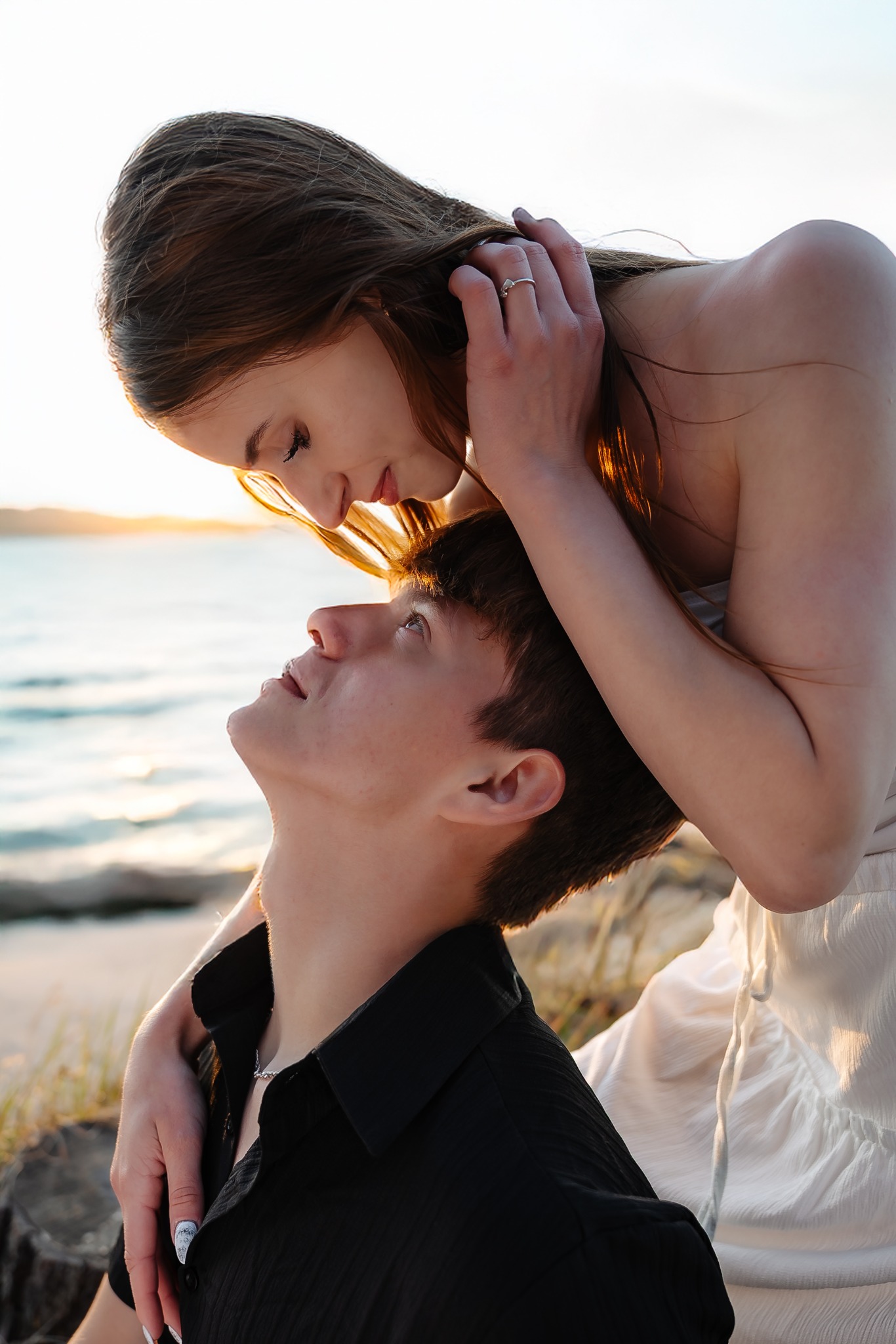 Zakochana para na plaży: kobieta w białej sukience obejmuje mężczyznę w czarnej koszuli, patrzącego w górę. Delikatne światło zachodzącego słońca.