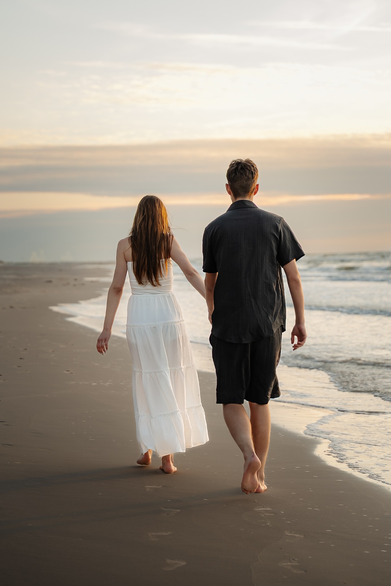 Para spaceruje brzegiem morza o zachodzie słońca, trzymając się za ręce. Kobieta w zwiewnej białej sukience, mężczyzna w ciemnym stroju. Widok z tyłu, boso po piasku.