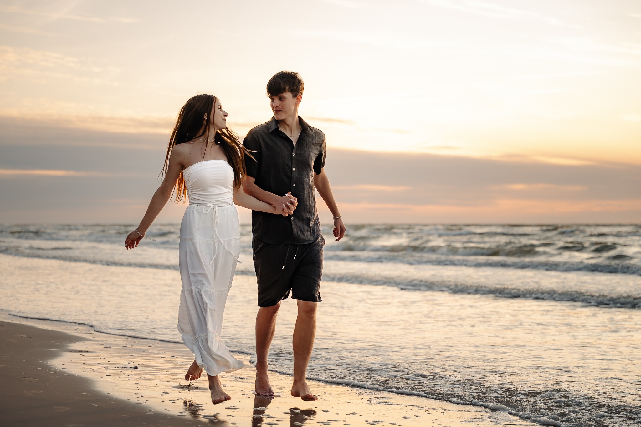 Zakochana para idzie brzegiem morza o zachodzie słońca. Kobieta w białej sukience, mężczyzna w czarnej koszuli i spodenkach trzymają się za ręce, patrząc sobie w oczy.