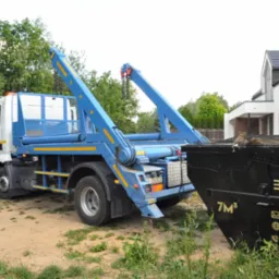 Ciężarówka hakowa z niebieskim dźwigiem i czarnym kontenerem na tle zieleni i nowoczesnego domu. Widoczny numer rejestracyjny pojazdu.