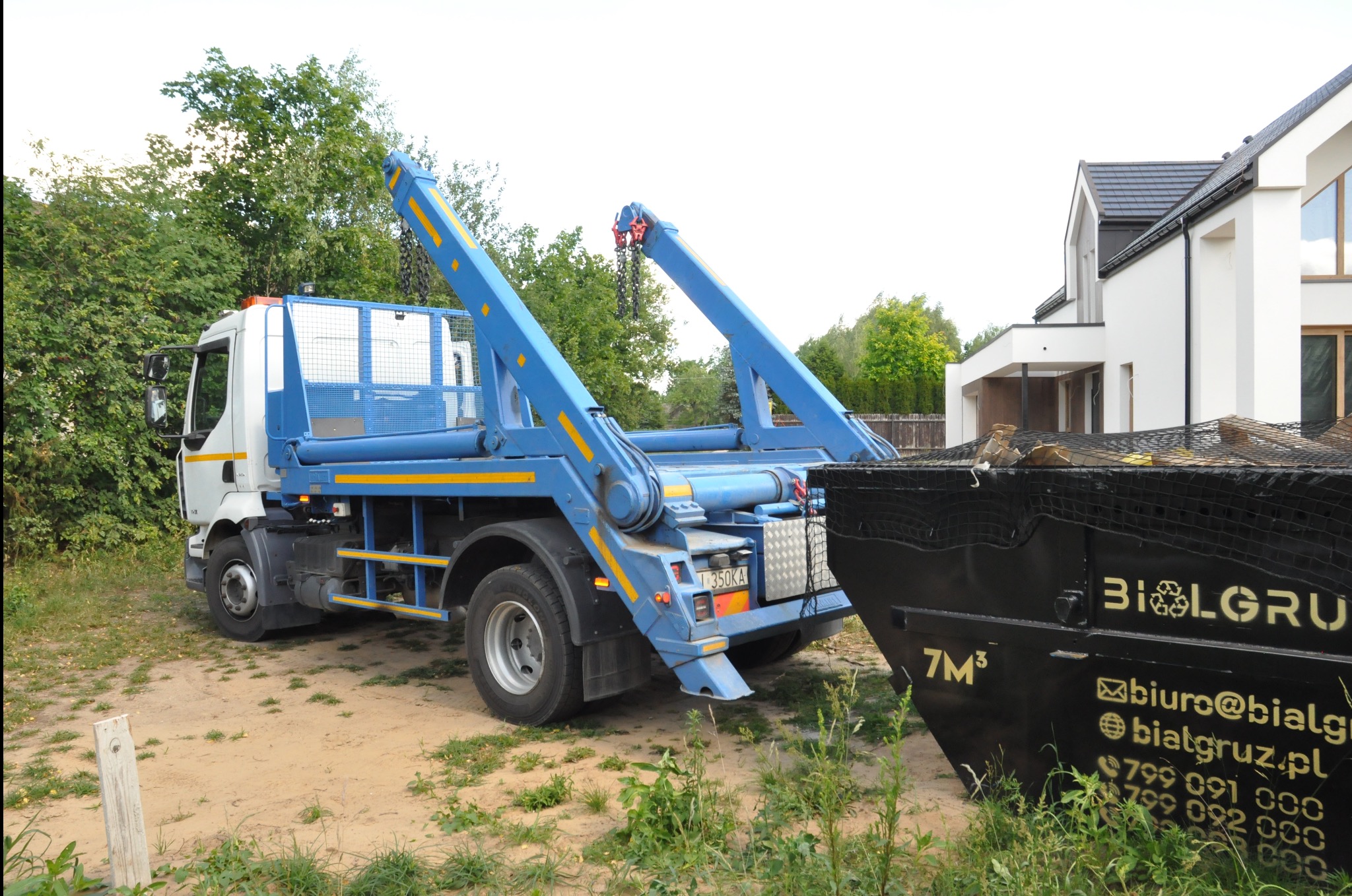 Ciężarówka hakowa z niebieskim dźwigiem i czarnym kontenerem na tle zieleni i nowoczesnego domu. Widoczny numer rejestracyjny pojazdu.