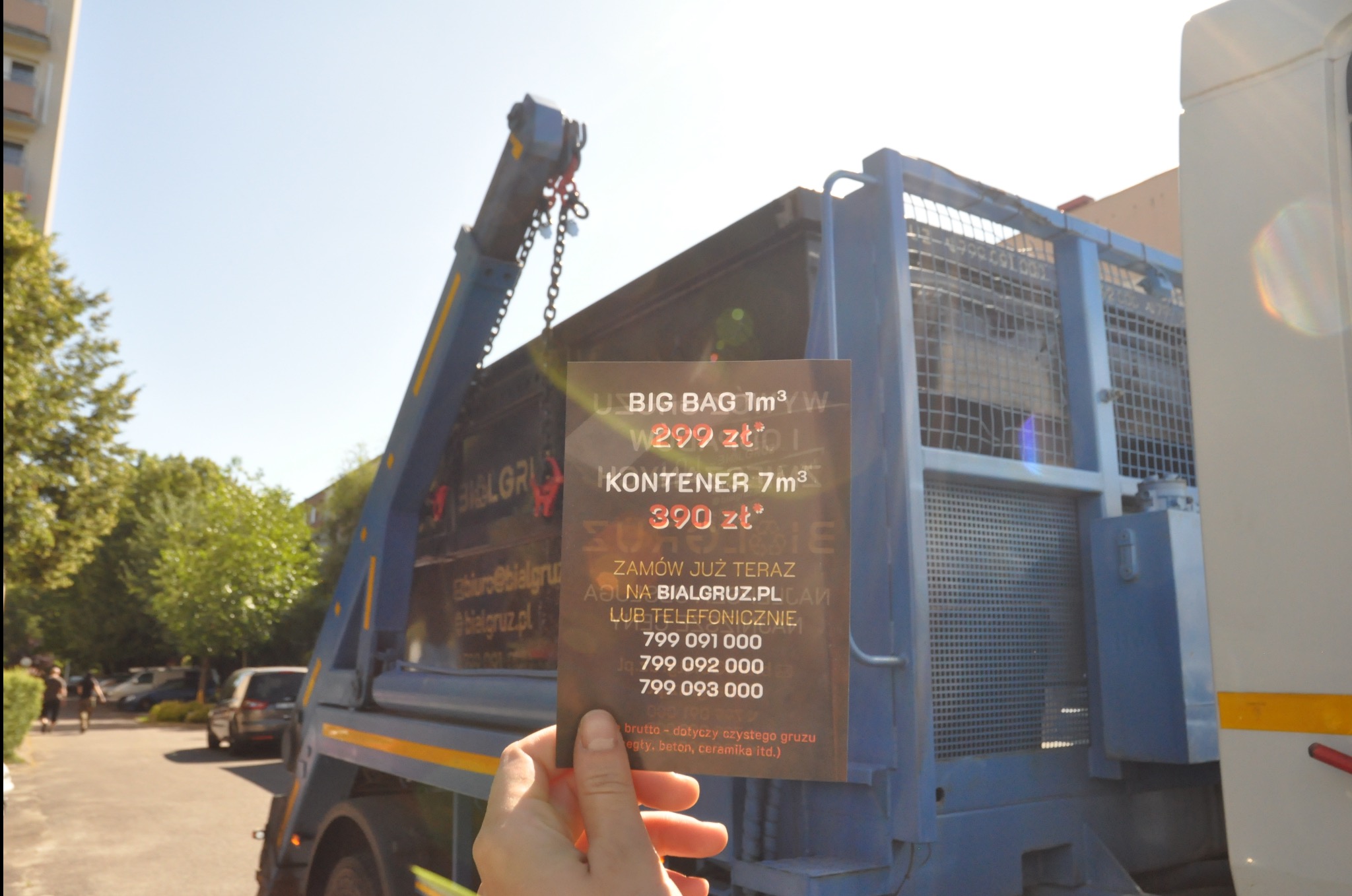 Ciężarówka z kontenerem na gruz w tle, w dłoni ulotka z ofertą wynajmu kontenerów i worków Big Bag. Reklama usług wywozu gruzu na tle pojazdu.