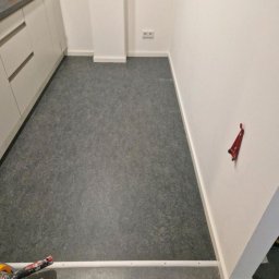 ML - Ukończona podłoga z szarego linoleum w Dębicy, widok z góry. Częściowo widoczne białe szafki kuchenne i narzędzia glazurnicze w rogu.