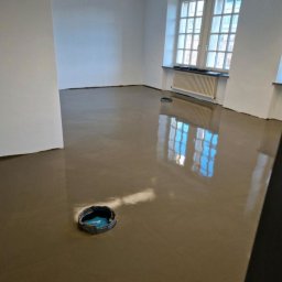 ML - Wnętrze po wylaniu samopoziomującej wylewki. Widoczne otwory instalacyjne i refleksy okien w gładkiej powierzchni podłogi. Minimalistyczny, nowoczesny styl.