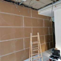 ML - Montaż płyt kartonowo-gipsowych na ścianie z widoczną drabiną i instalacją elektryczną pod sufitem. Etap wykończeniowy w pomieszczeniu.