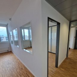 ML - Wnętrze biura po malowaniu: białe ściany, drewniana podłoga, okna z widokiem na miasto. Widoczny fragment puszki z farbą.