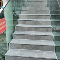 ML - Wykończone schody z szarych płytek z subtelnym wzorem, zabezpieczone szklaną balustradą z metalowymi poręczami. Widok z dołu, lekko z boku.