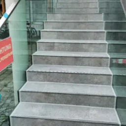 Wykończone schody z szarych płytek z subtelnym wzorem, zabezpieczone szklaną balustradą z metalowymi poręczami. Widok z dołu, lekko z boku.